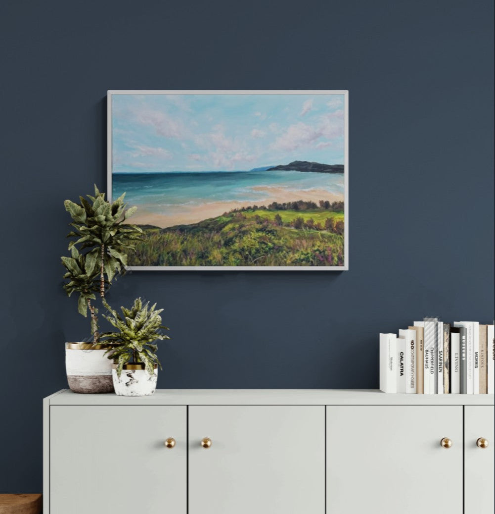 Expressive landscape painting of a calm beach viewed from a grassy headland. Hanging above a light gray cabinet with gold handles against a dark blue wall. Artwork by Judy Century Art.