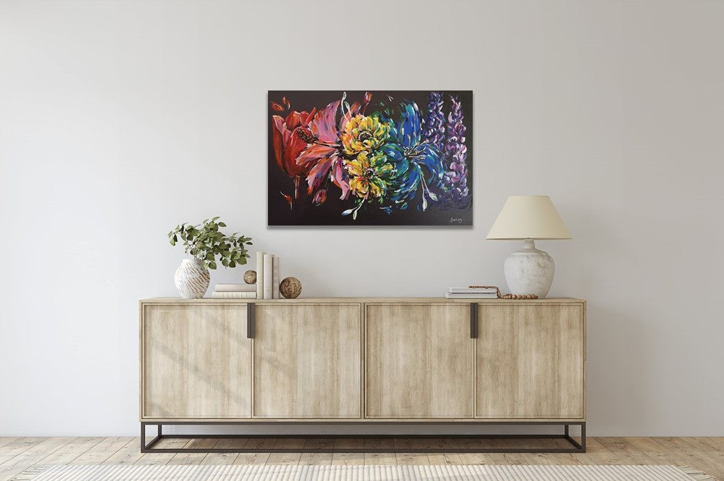 Rainbow flower painting on contrasting black background by Judy Century Art. Vibrant acrylic canvas hung on white wall above light wood side board with lamp and green eucalyptus leaves