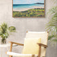 Framed original artwork for sale of a beach scene above a chair in a room with plants.