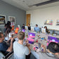 Group of people sitting around an office table with art supplies, engaged in a painting activity as a team building event with Judy Century Art.