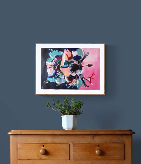 Original paper acrylic painting by Judy Century Art. Hibiscus Sorbet in black frame hanging on a navy wall above a wooden sideboard with plant in gold pot and wooden chair. Pinks, blues, teal, navy peach and white colours in elegant graphic interpretation. Shown framed in Wooden frame with white mount above wooden side board on navy wall.