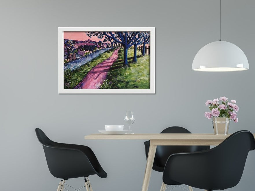 Colourful abstract landscape of an enchanting woodland scene with a shady path, trees, a river and colourful abstract bushes. Acrylic painting by Judy Century Art framed in white hanging above a dining table with black chairs