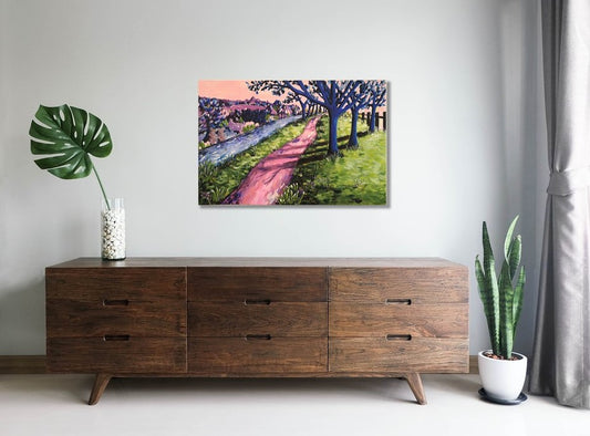 Colourful abstract landscape of an enchanting woodland scene with a shady path, trees, a river and colourful abstract bushes. Acrylic painting by Judy Century Art hanging in situ above a wooden sideboard with cheese plant leaf and plant